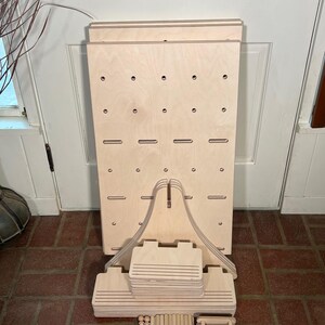 May include: A disassembled wooden display stand with a light wood finish. The stand features a large, rectangular backboard with evenly spaced holes and slots. Additional components include support pieces, dowels, and smaller wooden elements, all in a natural wood tone.