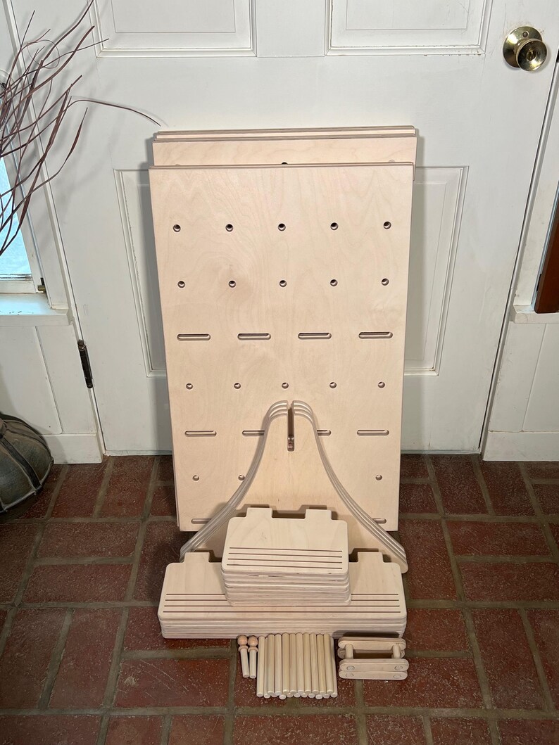 May include: A disassembled wooden display stand with a light wood finish. The stand features a large, rectangular backboard with evenly spaced holes and slots. Additional components include support pieces, dowels, and smaller wooden elements, all in a natural wood tone.