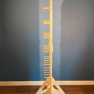 May include: A tall, light-colored wooden display stand featuring a central vertical spine with multiple horizontal pegs extending outward. The structure is supported by two triangular, cut-out base pieces that provide stability for the upright unit.