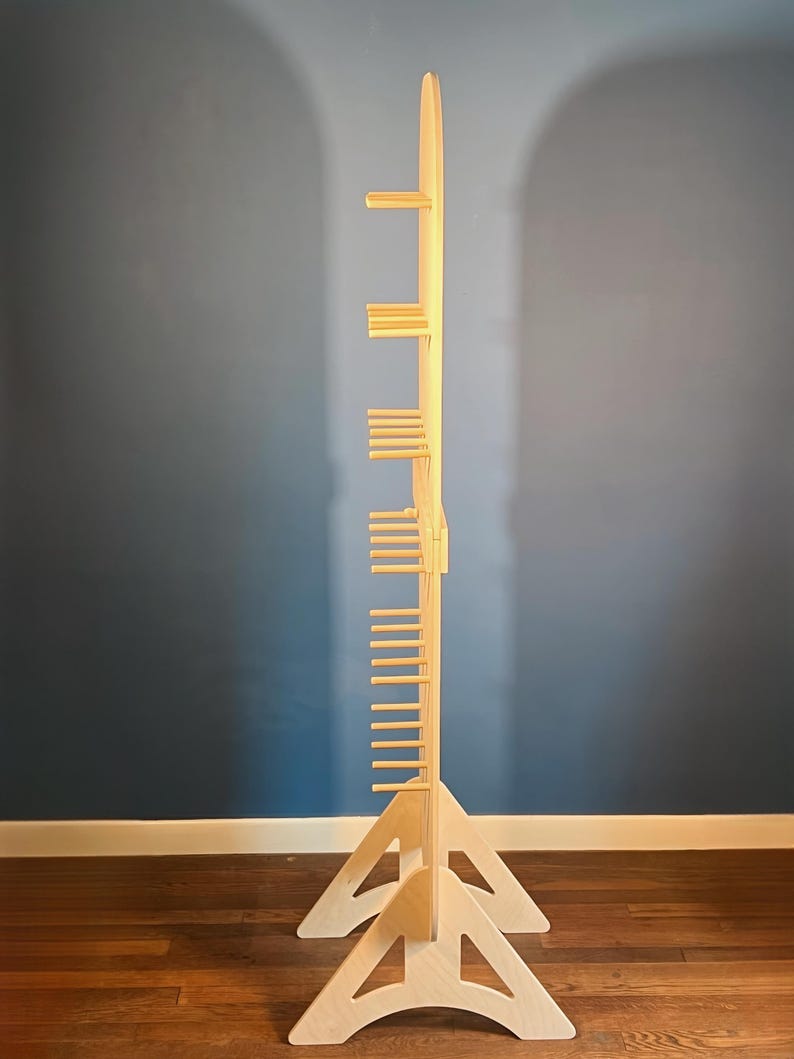 May include: A tall, light-colored wooden display stand featuring a central vertical spine with multiple horizontal pegs extending outward. The structure is supported by two triangular, cut-out base pieces that provide stability for the upright unit.