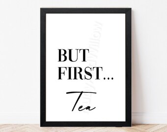 May include: A framed print with the text "BUT FIRST... Tea" in black against a white background. The print is in a black frame and is displayed on a white wall above a wooden floor.