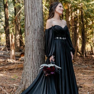 Peut inclure: Un couple dans un cadre forestier. La personne de gauche porte une robe noire à épaules dénudées avec une longue traîne et tient un bouquet. La personne de droite porte un costume noir avec un gilet vert. La scène est un mariage.