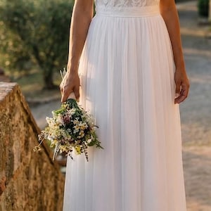Peut inclure: Une robe blanche longue jusqu'au sol avec un corsage en dentelle à manches courtes, une encolure ronde et une jupe fluide de couleur unie. Le vêtement est accompagné d'un petit bouquet de fleurs sauvages et de feuillage.