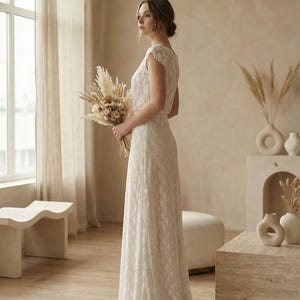 May include: A floor-length ivory dress featuring a delicate lace overlay with a repeating diamond pattern, short cap sleeves, and a fitted bodice that flows into a straight skirt. The garment is paired with a bouquet of dried pampas grass and flowers.