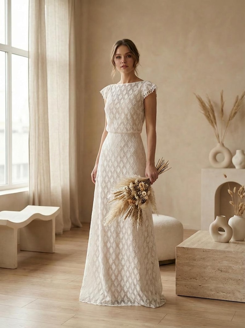 May include: A floor-length white dress featuring a repeating diamond pattern throughout the fabric, a boat neckline, and short cap sleeves with delicate lace trim. The garment has a fitted bodice that transitions into a flowing, straight skirt.