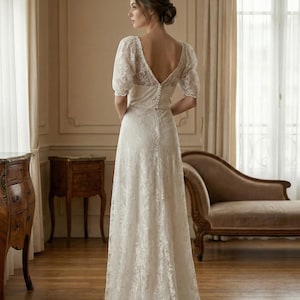 Pu&ograve; includere: Abito da sposa in pizzo bianco con schiena scoperta e maniche corte. L'abito ha una chiusura con bottoni sulla schiena e una gonna fluida. La sposa &egrave; in una stanza con mobili antichi e grandi finestre.