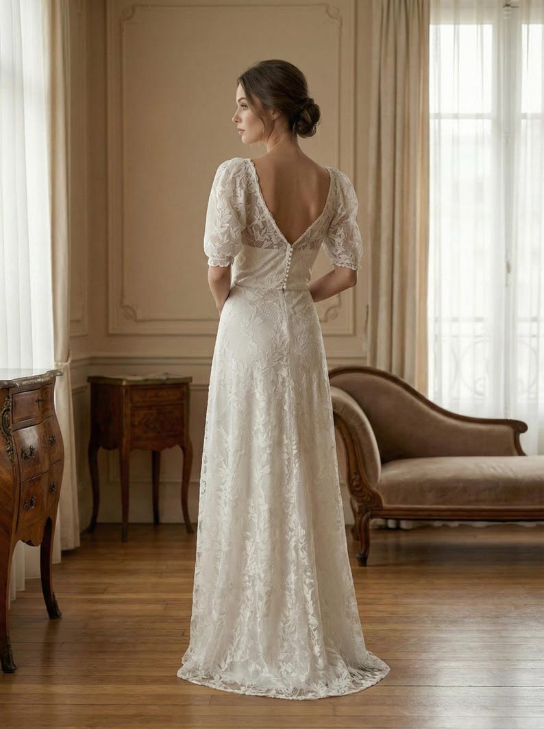Pu&ograve; includere: Abito da sposa in pizzo bianco con schiena scoperta e maniche corte. L'abito ha una chiusura con bottoni sulla schiena e una gonna fluida. La sposa &egrave; in una stanza con mobili antichi e grandi finestre.