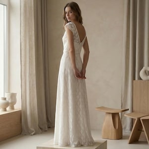 May include: A floor-length white lace dress featuring a V-neckline, cap sleeves, and a row of small buttons running down the back. The fabric has a repeating geometric pattern throughout, and the skirt falls in a straight silhouette to the floor.