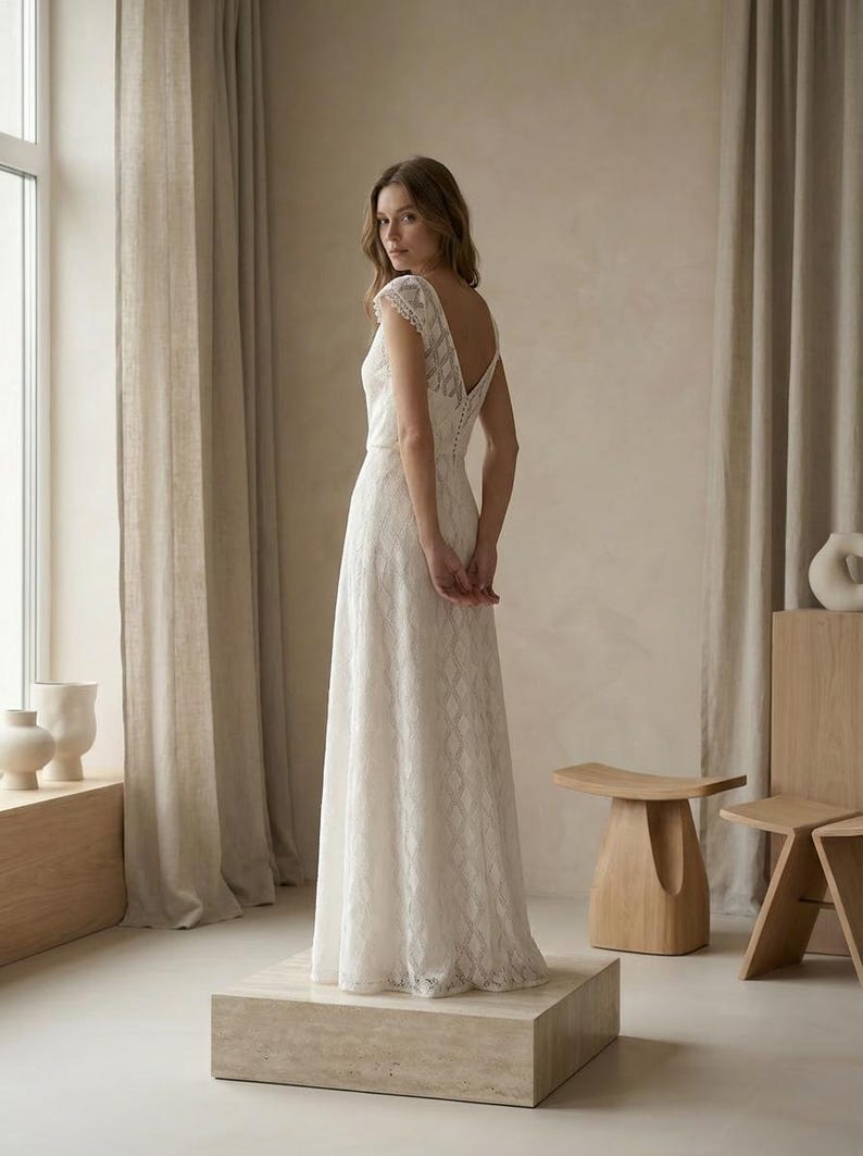 May include: A floor-length white lace dress featuring a V-neckline, cap sleeves, and a row of small buttons running down the back. The fabric has a repeating geometric pattern throughout, and the skirt falls in a straight silhouette to the floor.
