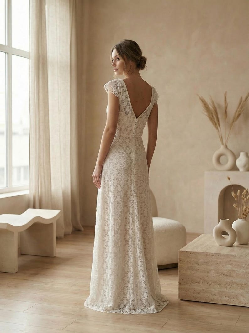 May include: A floor-length ivory lace dress featuring a geometric diamond pattern, short cap sleeves, and a deep V-neckline on the back. The bodice includes a vertical row of small fabric-covered buttons running down the center of the back.
