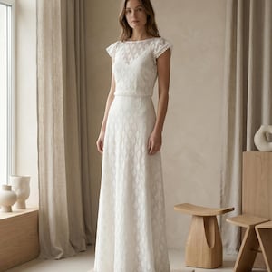 May include: A floor-length white dress featuring a diamond-shaped textured pattern throughout the fabric, short cap sleeves with delicate trim, and a boat neckline. The garment has a fitted bodice and a flowing skirt that reaches the floor.