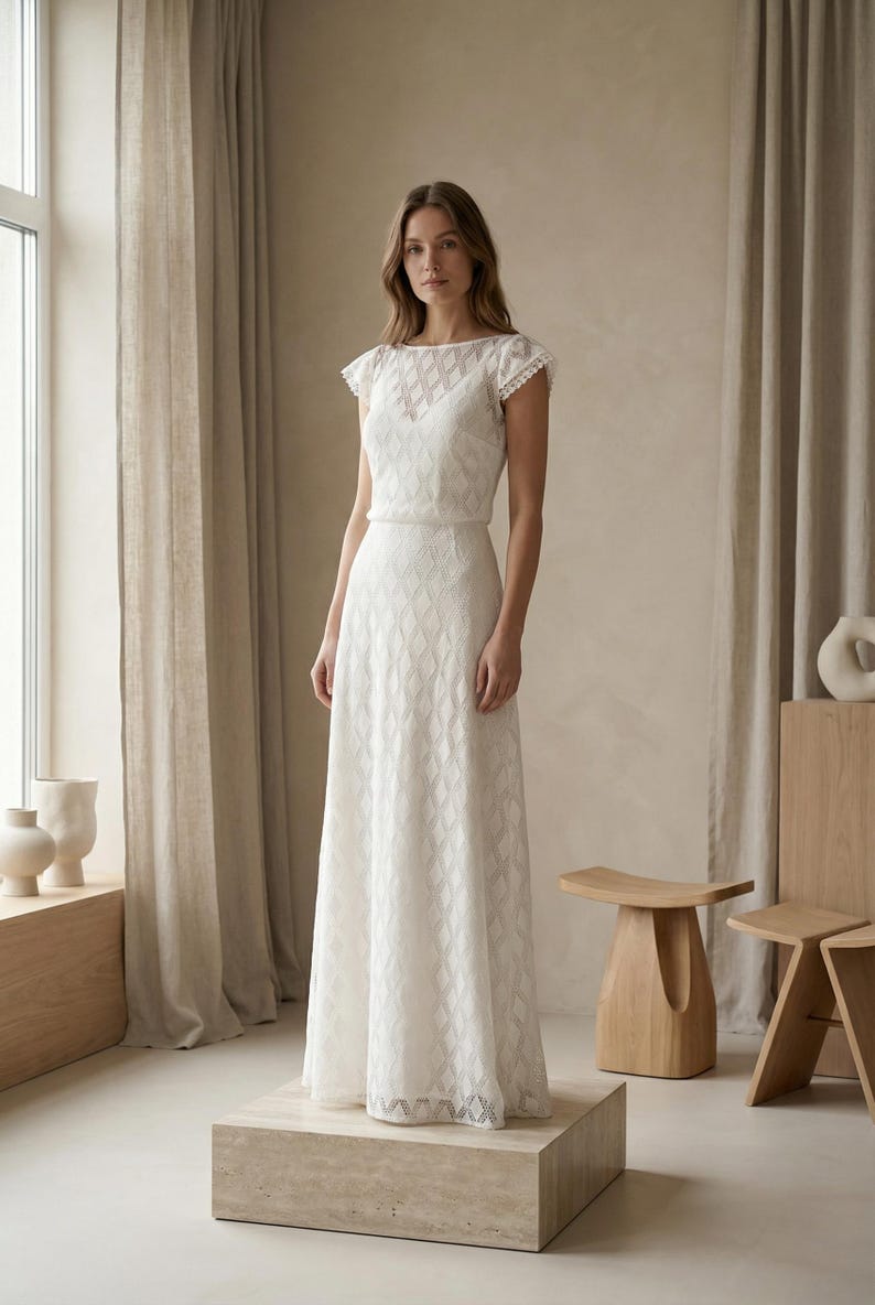 May include: A floor-length white dress featuring a diamond-shaped textured pattern throughout the fabric, short cap sleeves with delicate trim, and a boat neckline. The garment has a fitted bodice and a flowing skirt that reaches the floor.