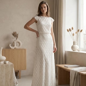 May include: A floor-length white dress featuring a geometric diamond pattern throughout the fabric, a boat neckline, and short cap sleeves with delicate scalloped edges. The garment has a fitted bodice and a straight, flowing skirt silhouette.