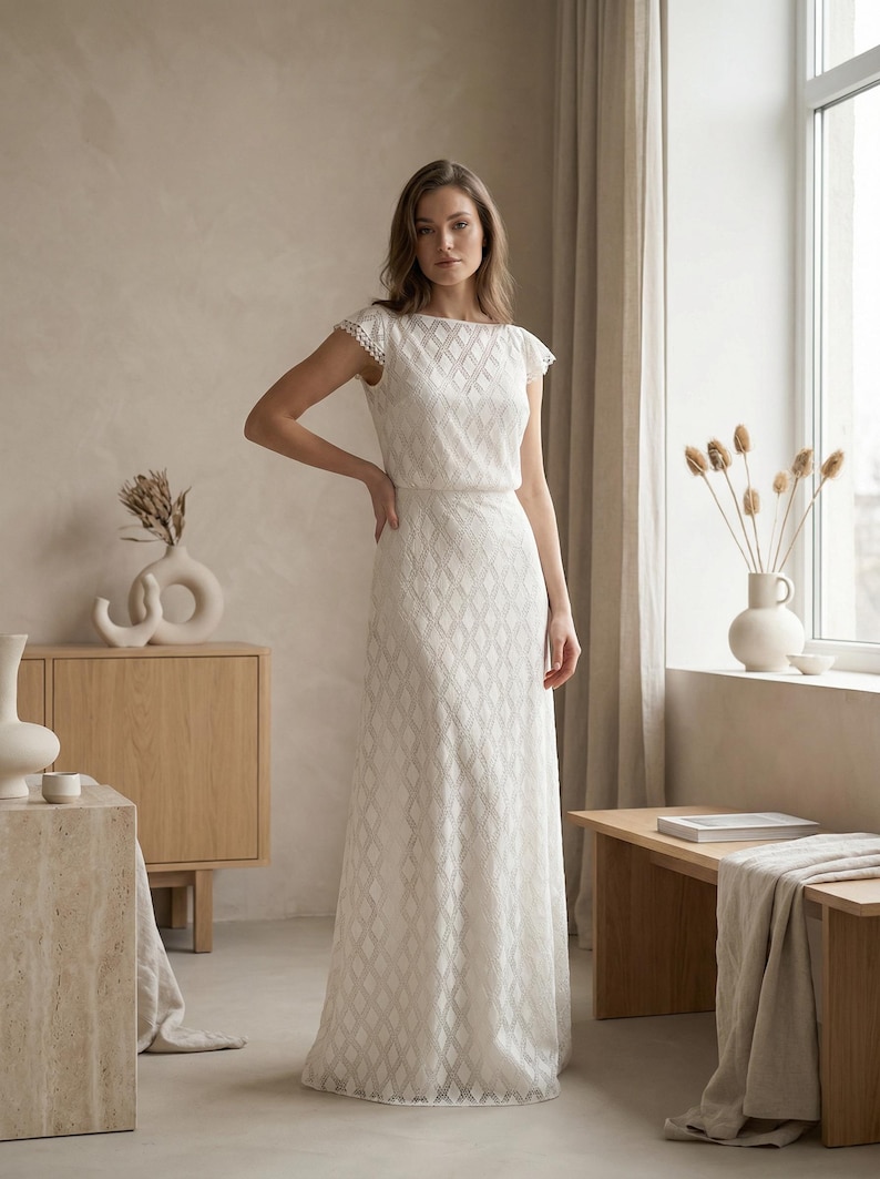 May include: A floor-length white dress featuring a geometric diamond pattern throughout the fabric, a boat neckline, and short cap sleeves with delicate scalloped edges. The garment has a fitted bodice and a straight, flowing skirt silhouette.