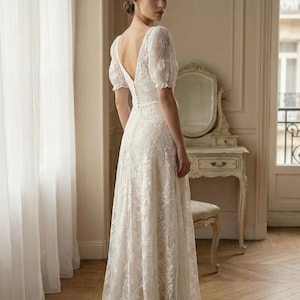 Pu&ograve; includere: Abito da sposa in pizzo bianco con scollo a V e maniche corte a sbuffo. L'abito presenta una schiena scoperta e una gonna fluida. La sposa &egrave; in una stanza con una finestra e una toeletta.