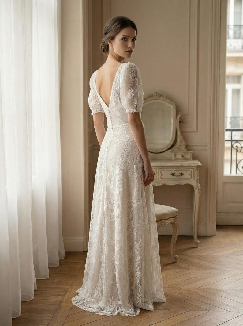 Pu&ograve; includere: Abito da sposa in pizzo bianco con scollo a V e maniche corte a sbuffo. L'abito presenta una schiena scoperta e una gonna fluida. La sposa &egrave; in una stanza con una finestra e una toeletta.