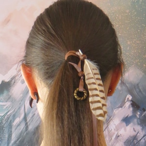 May include: A person with long brown hair tied back with a brown leather cord and a feather accent. The hair tie has a small gold charm. The person is wearing a brown hooded top.