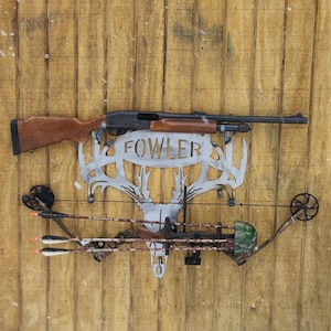 Peut inclure: Une décoration murale en métal représentant un fusil de chasse et un arc et des flèches, montée sur un mur en bois. Le fusil de chasse a une crosse en bois marron et la pièce d'art en métal porte le nom "FOWLER". L'arc et les flèches ont des accents de camouflage.