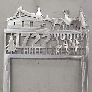 Op de afbeelding: Een metalen tuinschild met een silhouet van een huis, bomen en herten. Het bord staat "1722 Woody Lane Three Lakes WI".
