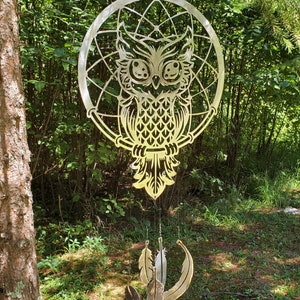 May include: A metal dream catcher with an owl design and feathers hanging from the bottom. The dream catcher is gold in color and is hanging from a tree branch.