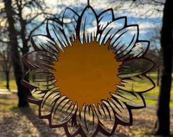 May include: A metal sunflower-shaped ornament with a yellow center and brown petals hanging from a string in front of a blurred background of trees and a blue sky.