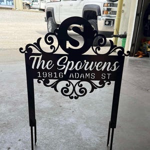 Könnte beinhalten: Schwarzes Metall-Gartenschild mit dem Namen "The Sporwens", der Adresse "19816 ADAMS ST" und einem großen "S" in einem Kreis. Das Schild hat dekorative Schnörkel und zwei Pfähle zur Befestigung im Boden.