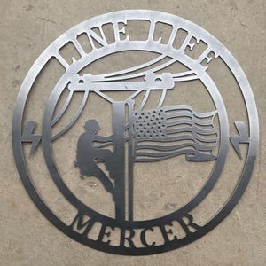 Puede incluir: Un letrero redondo de metal con la silueta de un lineman subiendo a un poste de electricidad con una bandera estadounidense. El letrero dice "One Life Mercer".
