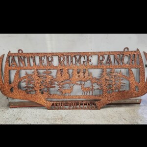 May include: A rusty metal sign with the words "Antler Ridge Ranch" cut out. The sign features a silhouette of two deer in a forest setting.