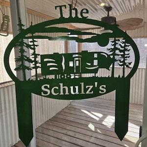 May include: Green metal sign with an oval shape featuring a silhouette of a camper, trees, and a moon. The text "The Schulz's" is written below the camper.