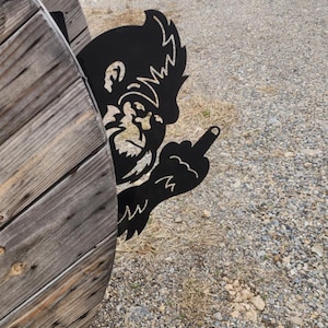 May include: A black metal silhouette of a cartoon bear with a mischievous expression, giving the middle finger, is attached to a wooden spool. The bear is partially obscured by the spool, and the background is a gravel road.