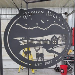 May include: Black metal sign with a cut-out design of a moon, stars, mountains, a barn with the text "Johnson" on it, a willow tree, a deer, and a smaller deer. The sign reads "Hidden Hills Est. 1987".