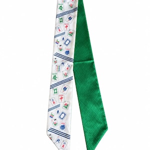 May include: A long, narrow scarf featuring a white side with a repeating pattern of colorful Mahjong tiles and blue diagonal stripes, and a solid green side with a subtle, textured chevron pattern throughout the fabric surface.