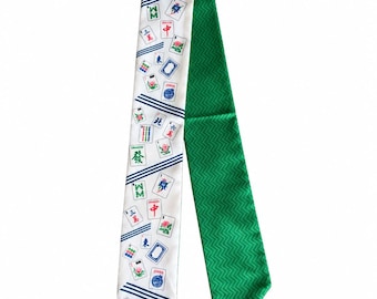 May include: A long, narrow scarf featuring a white side with a repeating pattern of colourful Mahjong tiles and blue diagonal stripes, and a solid green side with a subtle, textured chevron pattern throughout the fabric surface.