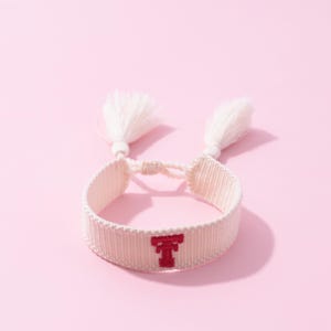 May include: A woven bracelet in a light pink hue, featuring a red embroidered letter "T" in the center. The bracelet is adorned with two white tassel accents on each end. The background is a solid pink.