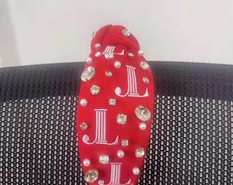 May include: Red fabric headband with a knotted design and white embroidered letters "JL" on the front. The headband is decorated with clear and pearl-like rhinestones.