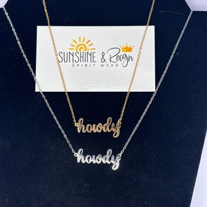 May include: Two gold necklaces with the word "howdy" written in cursive on them. The necklaces are on a black display stand. The necklaces are from the company "Sunshine & Reign Spirit Wear."