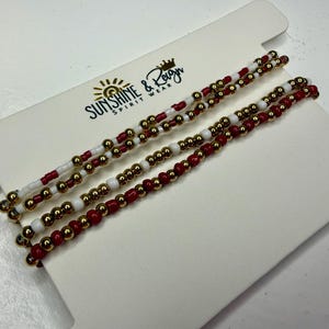 May include: Three beaded necklaces with red, white, and gold beads are displayed on a white card. The necklaces are arranged in parallel lines, showcasing the alternating color patterns. The card has the text "Sunshine & Reign Spirit Wear".
