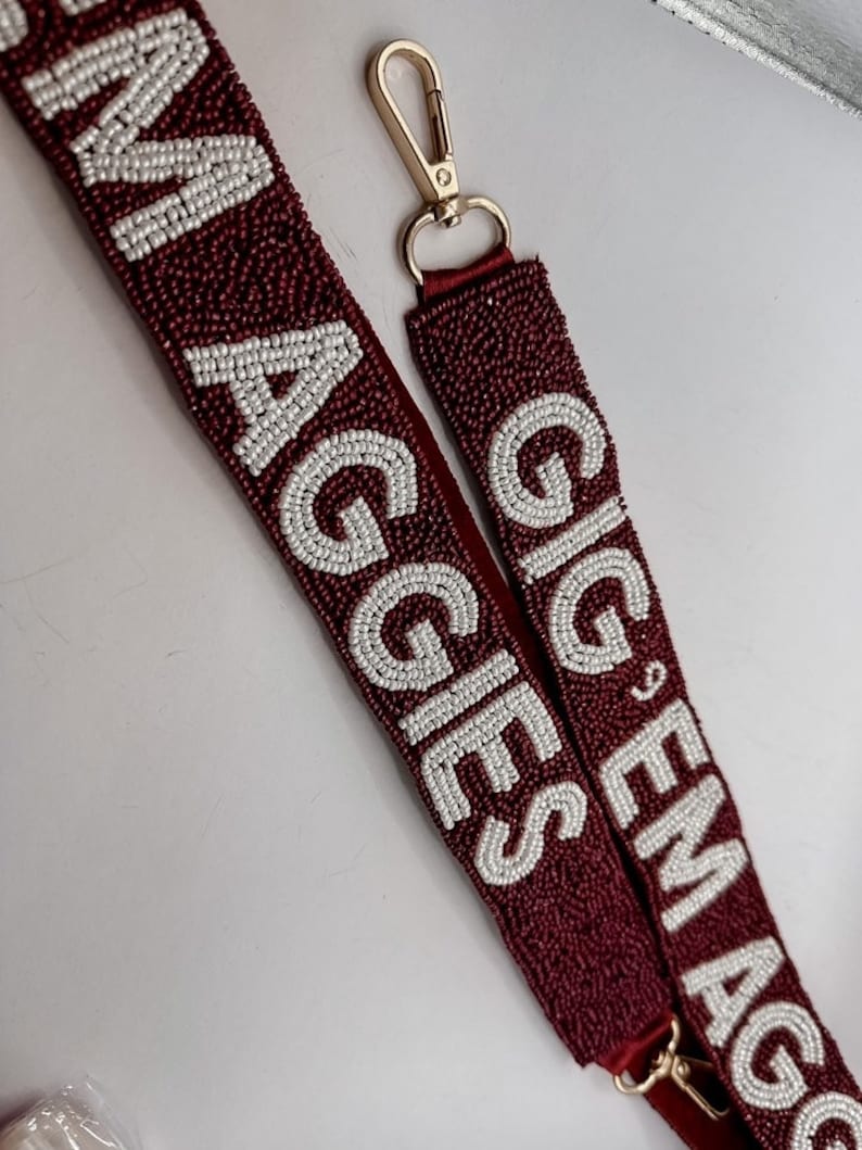 May include: A maroon beaded bag strap with white beads spelling out "Gig 'Em Aggies". The strap has a gold clasp.