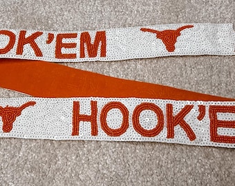 May include: A white strap with the words "HOOK 'EM" in orange beads, along with a longhorn symbol. The strap has silver-colored metal clasps on each end and an orange fabric backing.