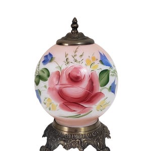 May include: A decorative, round lamp with a pink and white glass globe featuring a large, hand-painted red rose and floral accents. The lamp has a bronze-colored metal base and a matching finial on top.