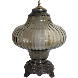 May include: A decorative glass lamp with a ribbed, olive-green shade and a bronze-colored base. The lamp has a bulbous shape with a lid and a decorative finial. The base features an ornate, floral design.