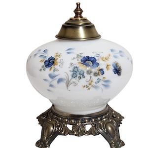 May include: White glass lamp with a brass lid and ornate base. The lamp features a floral design in blue and yellow. The base has a decorative, antique-style design. A vintage-style decorative lamp.