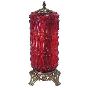 May include: A red glass jar with a geometric pattern, a decorative lid, and a bronze-colored base. The jar is cylindrical and stands on a three-footed base. The lid is also bronze-colored and has a small, ornate finial.