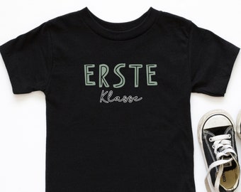 May include: A black short-sleeved t-shirt with the words "ERSTE Klasse" in light green. The shirt is paired with a pair of black and white sneakers with white laces. The text is in a sans-serif font with a cursive font for "Klasse".