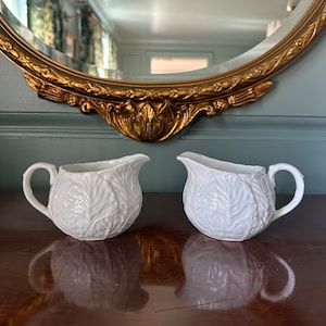 Puede incluir: Dos jarras de crema de cerámica blanca con un diseño de hojas. Las jarras están sobre una superficie de madera oscura.