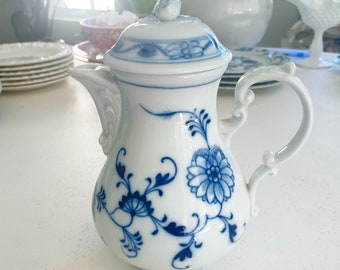 May include: A white porcelain creamer with a blue and white floral pattern. The creamer has a lid and a handle.