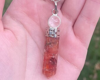 May include: A silver chain necklace with a clear quartz crystal pendant. The pendant is set in a silver bezel and has a silver filigree design. The crystal is a point shape and is a light orange color.