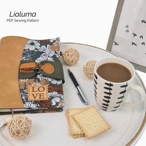 Könnte beinhalten: Ein brauner und grüner Blumenstoff-Notizbuchumschlag mit einem braunen Lederriemen und einer Schnalle. Der Umschlag trägt den Schriftzug "LOVE". Das Notizbuch liegt auf einem weißen Tablett mit einer Kaffeetasse, Keksen und Dekorationskugeln.