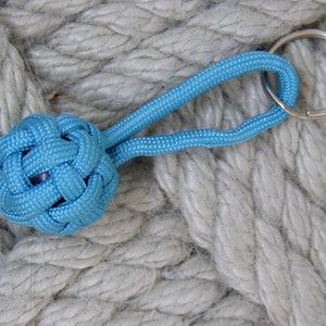 Peut inclure: Un porte-clés en corde bleue avec une boule nouée à l'extrémité. Le porte-clés est sur un fond de corde blanche.