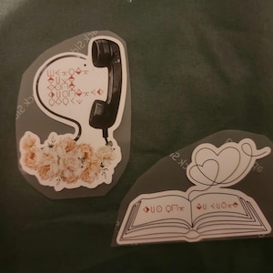 Op de afbeelding: Twee vinylstickers. Eén toont een zwarte vintage telefoonhoorn verbonden met een tros licht perzikkleurige bloemen. De andere toont een open boek met een doorlopende lijntekening van twee harten erboven, voorzien van geometrische symbolen.